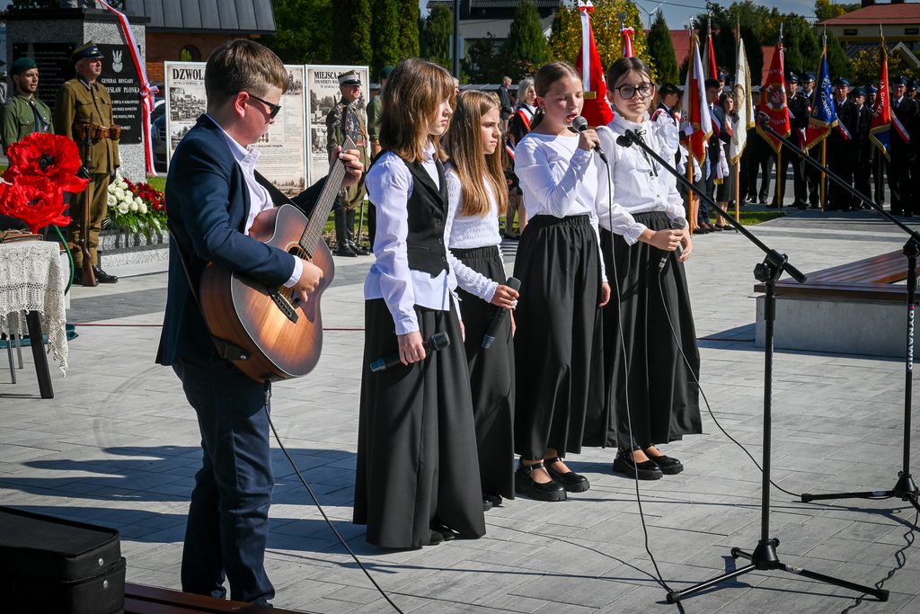 Grupa dzieci ubranych na biało i czarno, chłopiec gra na gitarze, dziewczynki trzymają mikrofony, w tle żołnierze, flagi i tablice informacyjne.