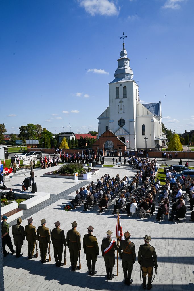 Grupa żołnierzy w mundurach na pierwszym planie, z tyłu ludzie na krzesłach, biały kościół z metalowym dachem, niebieskie niebo z kilkoma chmurami, teren z kwiatami i pomnikiem.