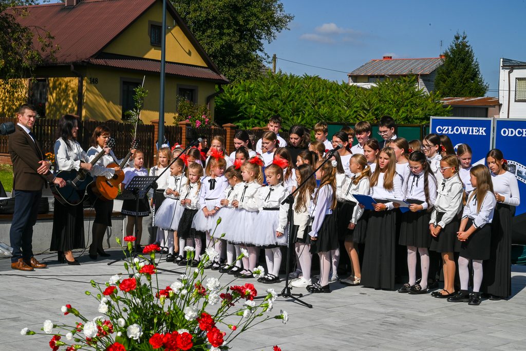 Grupa dzieci i dorosłych w białych bluzkach i czarnych spódnicach oraz spodniach na zewnątrz, dwie kobiety grają na gitarach, w tle zielone krzewy i żółty dom.