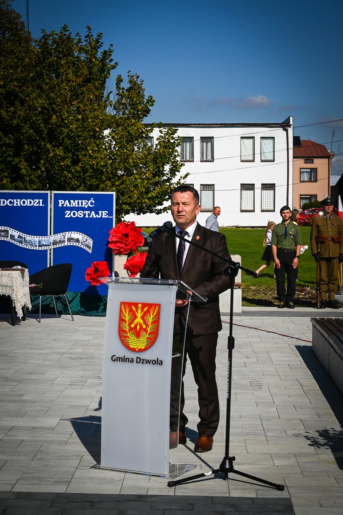 Mężczyzna w garniturze przemawia przy przezroczystym mównicy z herbem i napisem "Gmina Dzwola", w tle zielone drzewa, niebieskie plansze z napisem, ludzie w mundurach i białe budynki.