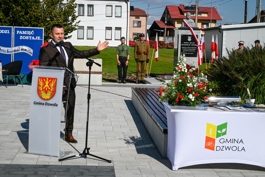 Mężczyzna w garniturze przy mównicy z herbem Gmina Dzwola, w tle pomnik z popiersiem, żołnierze w mundurach, biało-czerwone flagi i kwiaty.