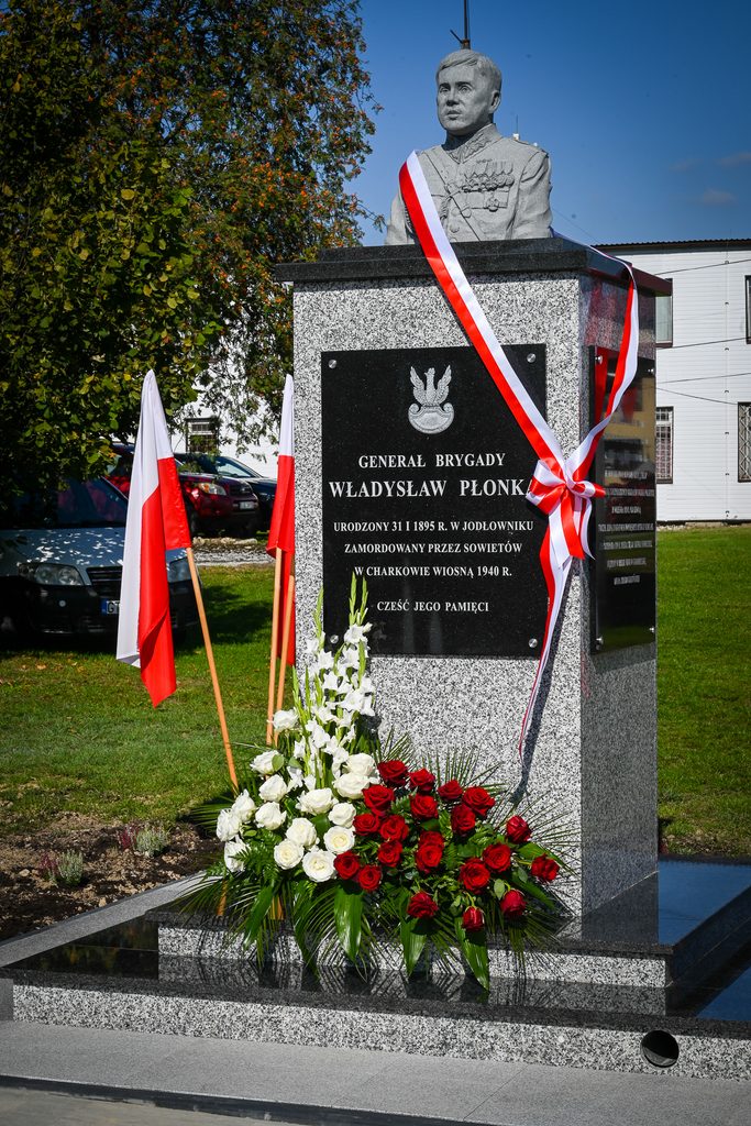 Popiersie mężczyzny w mundurze na szarej marmurowej podstawie z czarną tablicą, biało-czerwone wstążki, biało-czerwone flagi i bukiet białych i czerwonych kwiatów przed pomnikiem.