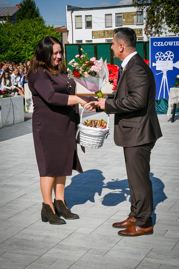 Mężczyzna w brązowym garniturze wręcza kobiecie w brązowej sukience bukiet kwiatów i kosz z cukierkami na tle grupy siedzących dzieci oraz budynku.