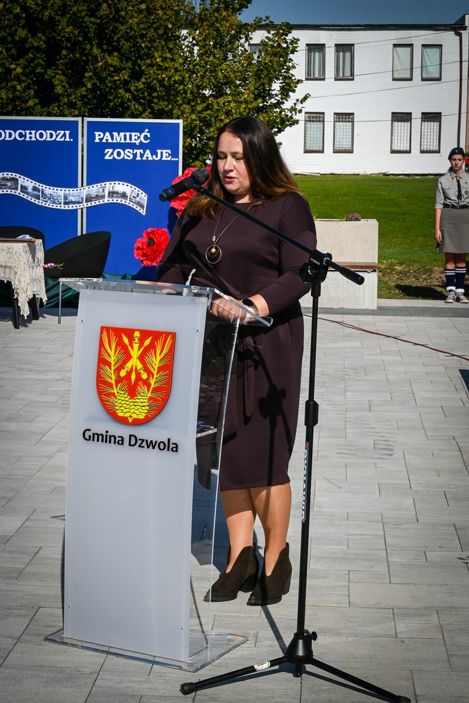 Kobieta w ciemnej sukience przemawia przy przezroczystej mównicy z herbem i napisem "Gmina Dzwola", za nią niebieskie plansze z napisem i zielone drzewo.