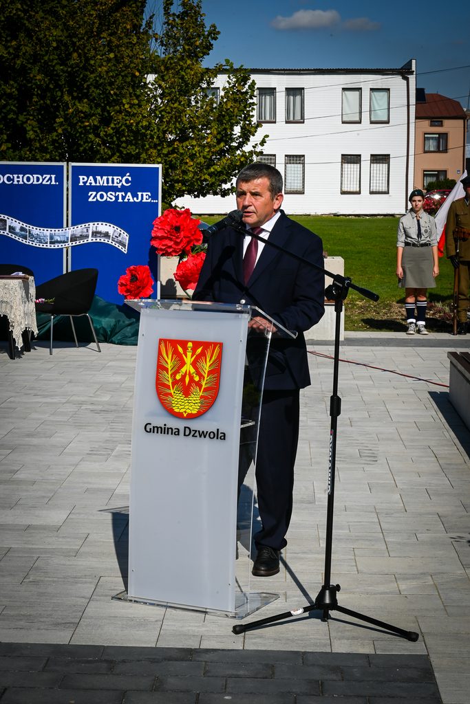Mężczyzna w ciemnym garniturze przemawia przy przezroczystym mównicy z herbem i napisem "Gmina Dzwola", w tle zielone drzewo, biały budynek, niebieskie tablice i dwie osoby w mundurach.