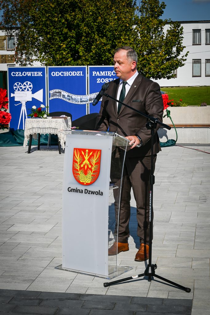 Mężczyzna w brązowym garniturze przemawia przy przezroczystym mównicy z herbem i napisem Gmina Dzwola, na tle zielonego drzewa i granatowych plansz z białym napisem.
