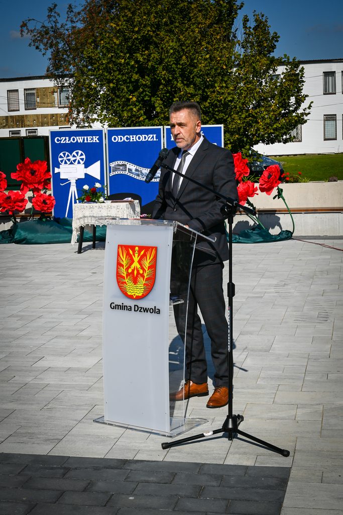 Mężczyzna w garniturze stoi za transparentnym mównicą z herbem i napisem Gmina Dzwola, obok mikrofon na statywie, w tle duże czerwone kwiaty i zielone drzewo.