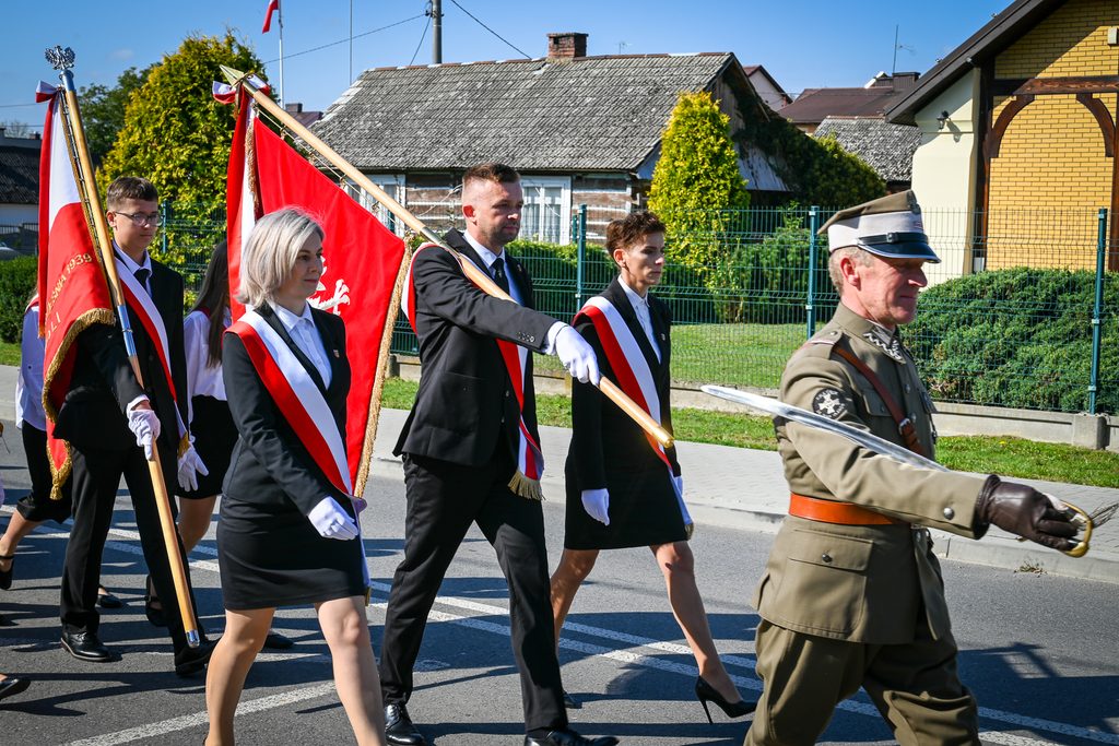Grupa osób w eleganckich strojach z białymi rękawiczkami, niosąca czerwone flagi z białymi orłami, na ulicy w słoneczny dzień, obok mężczyzna w mundurze wojskowym z szablą.