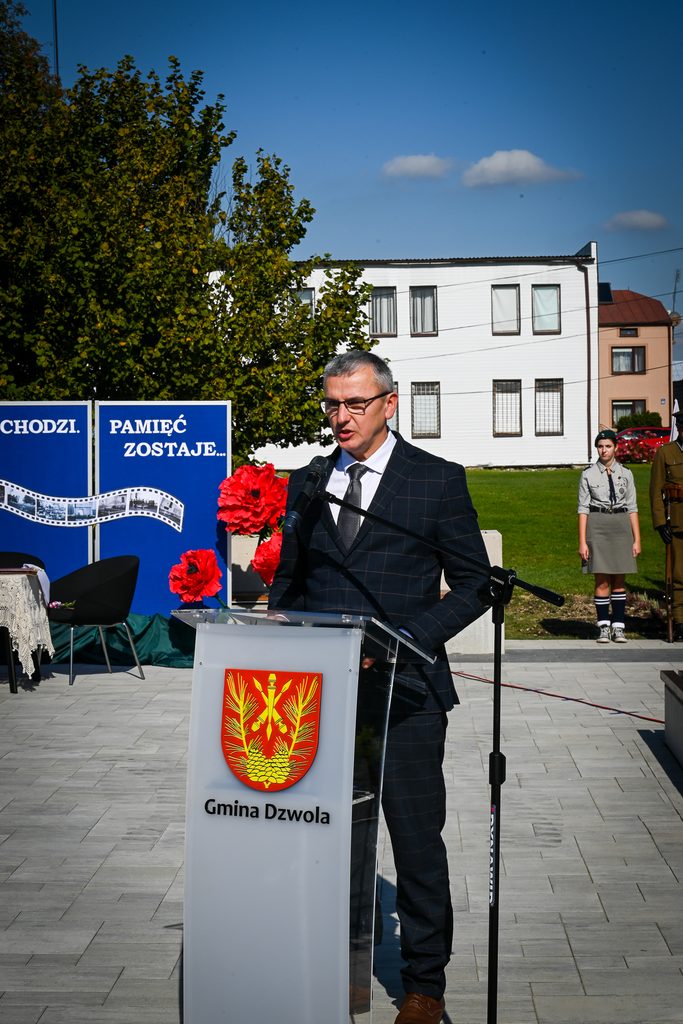 Mężczyzna w garniturze przemawia przy mównicy z herbem i napisem Gmina Dzwola, w tle duże czerwone kwiaty, zielone drzewo, budynki i dwie osoby w mundurach.