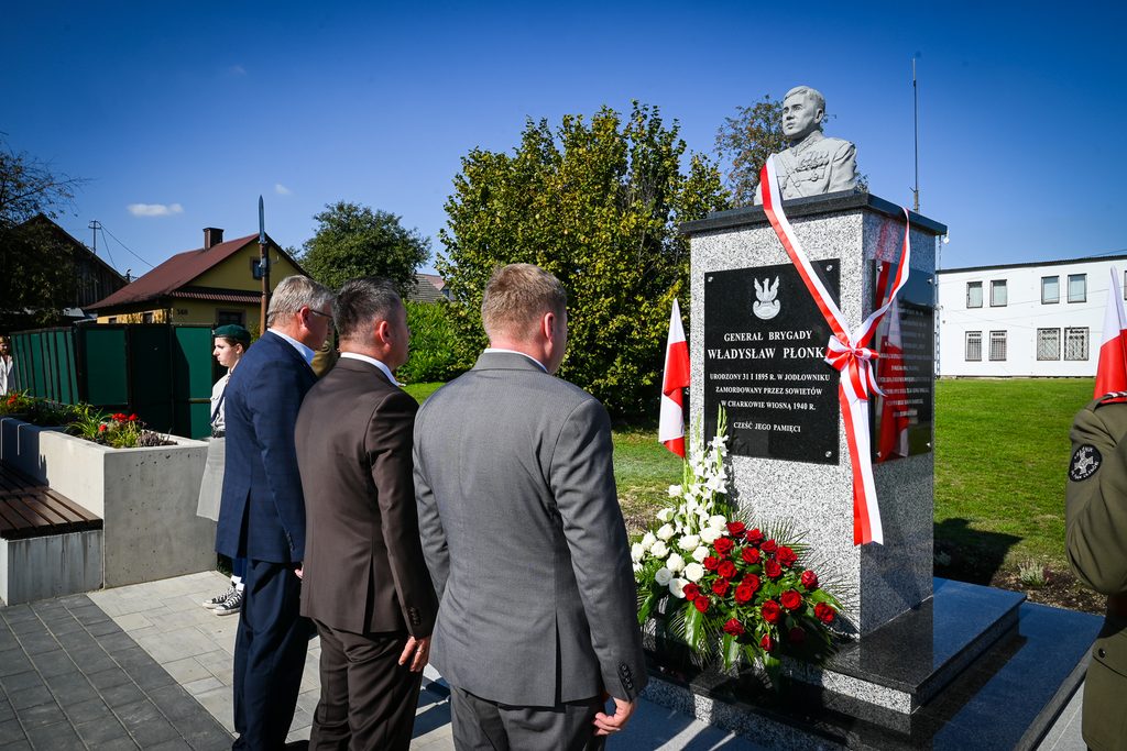 Pomnik generała Władysława Płonki z biało-czerwoną szarfą i wieńcem z czerwonych i białych róż, przed którym stoją trzy osoby w garniturach, na tle zieleni i budynków.