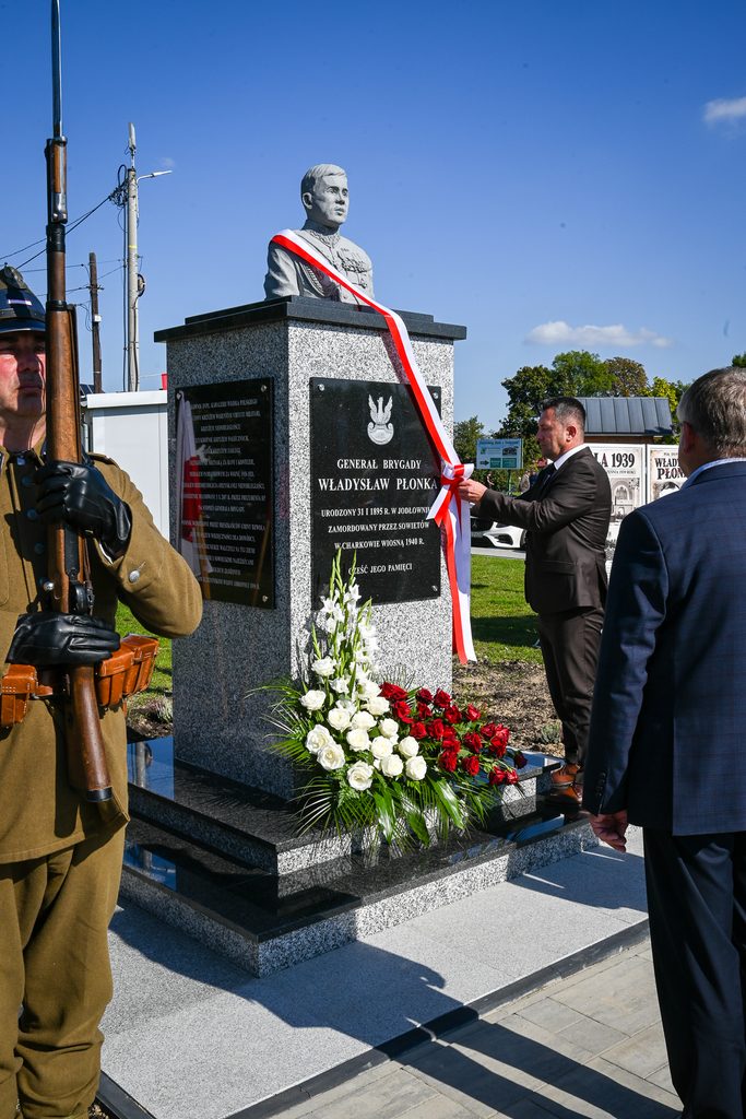 Pomnik generała Władysława Płonki z szarfą w barwach biało-czerwonych, kwiaty białe i czerwone, mężczyzna w garniturze, żołnierz w mundurze z karabinem, niebieskie niebo.