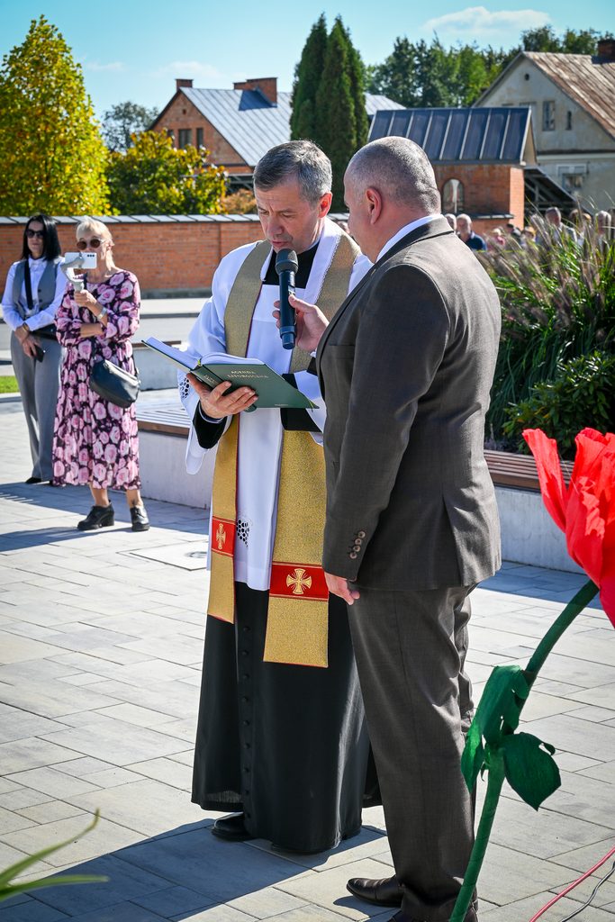 Mężczyzna w sutannie z białym stułą i księgą oraz mężczyzna w garniturze trzymający mikrofon, w tle kobiety i domy, na pierwszym planie duży czerwony kwiat.