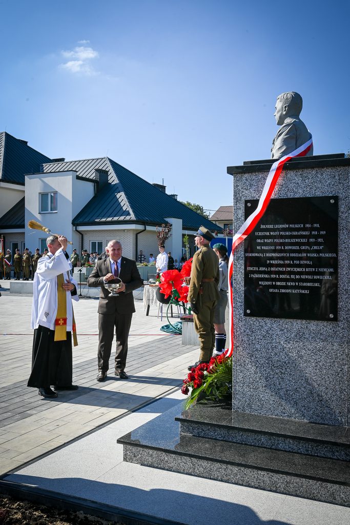 Pomnik z napisem upamiętniającym żołnierzy legionów polskich z biało-czerwoną szarfą, obok stoi ksiądz z kropidłem i mężczyzna w garniturze, w tle żołnierze i budynki.