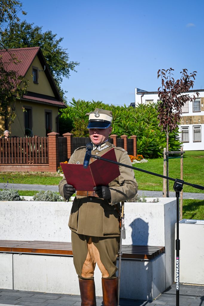 Mężczyzna w mundurze wojskowym, beżowy płaszcz i czapka, czyta z czerwonej teczki przed mikrofonem na tle zieleni, domów i niebieskiego nieba.