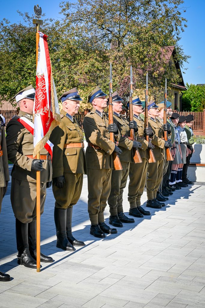 Mężczyźni w mundurach wojskowych z karabinami i proporcem, obok dzieci w mundurach harcerskich, stojący na chodniku, w tle drzewa i dom z czerwoną dachówką.