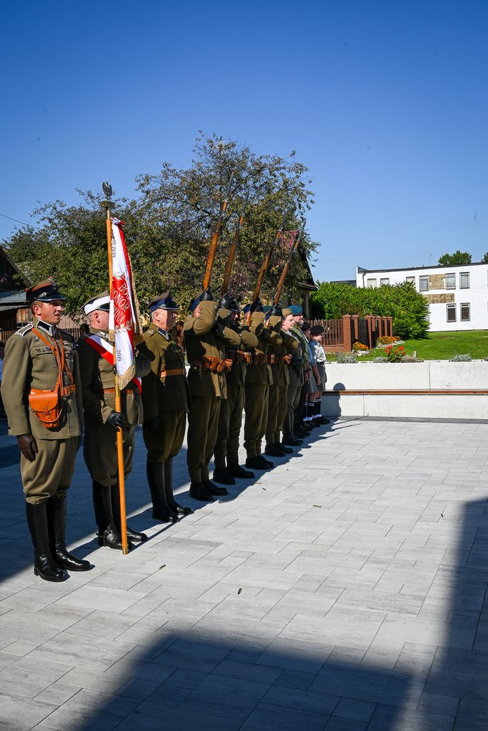 Grupa żołnierzy w mundurach wojskowych i harcerzy ustawionych w szeregu na jasnej kostce brukowej, w tle drzewa i budynki, pod jasnym niebem.