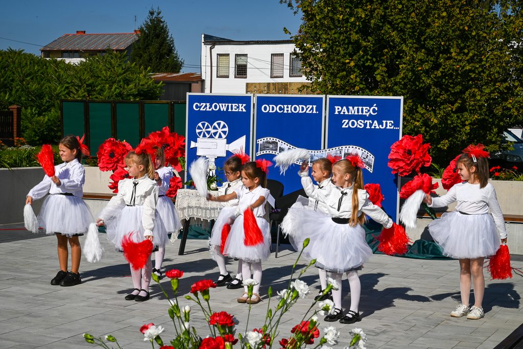 Grupa dziewczynek w białych strojach i czerwonych kokardach tańczy na tle niebieskich tablic z napisem "Człowiek odchodzi. Pamięć zostaje..." oraz dużych czerwonych kwiatów.