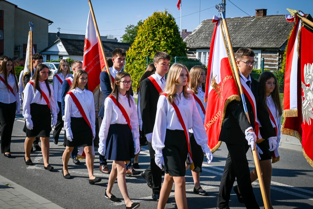 Grupa młodych ludzi w białych koszulach, czarnych spódnicach i spodniach, z czerwono-białymi szarfami i rękawiczkami, niosąca czerwono-białe flagi w marszu na ulicy.