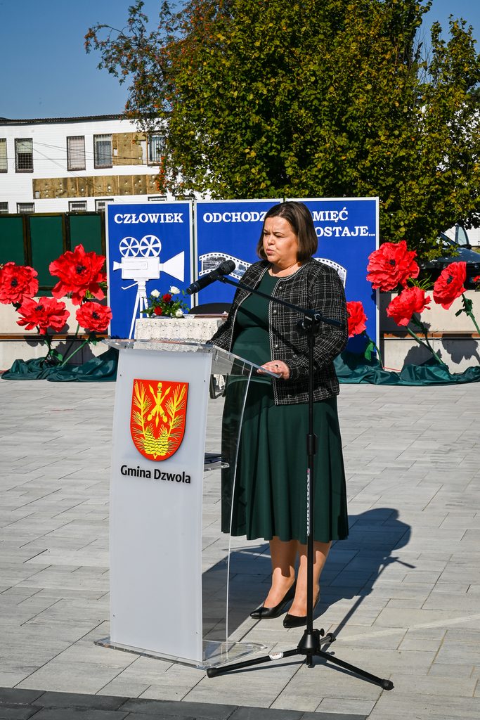 Kobieta w zielonej sukience i czarnej marynarce stoi przy przezroczystym mównicy z herbem i napisem Gmina Dzwola, z tyłu niebieskie tablice i czerwone dekoracyjne kwiaty.