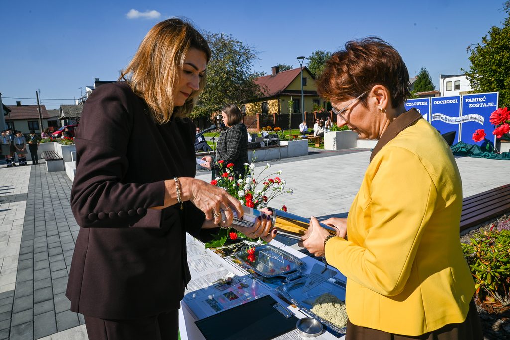 Dwie kobiety trzymają metalową tubę, stoją przy stole z dokumentami i czerwono-białymi kwiatami, w tle budynki, niebieskie niebo, ludzie i plansza z napisem.