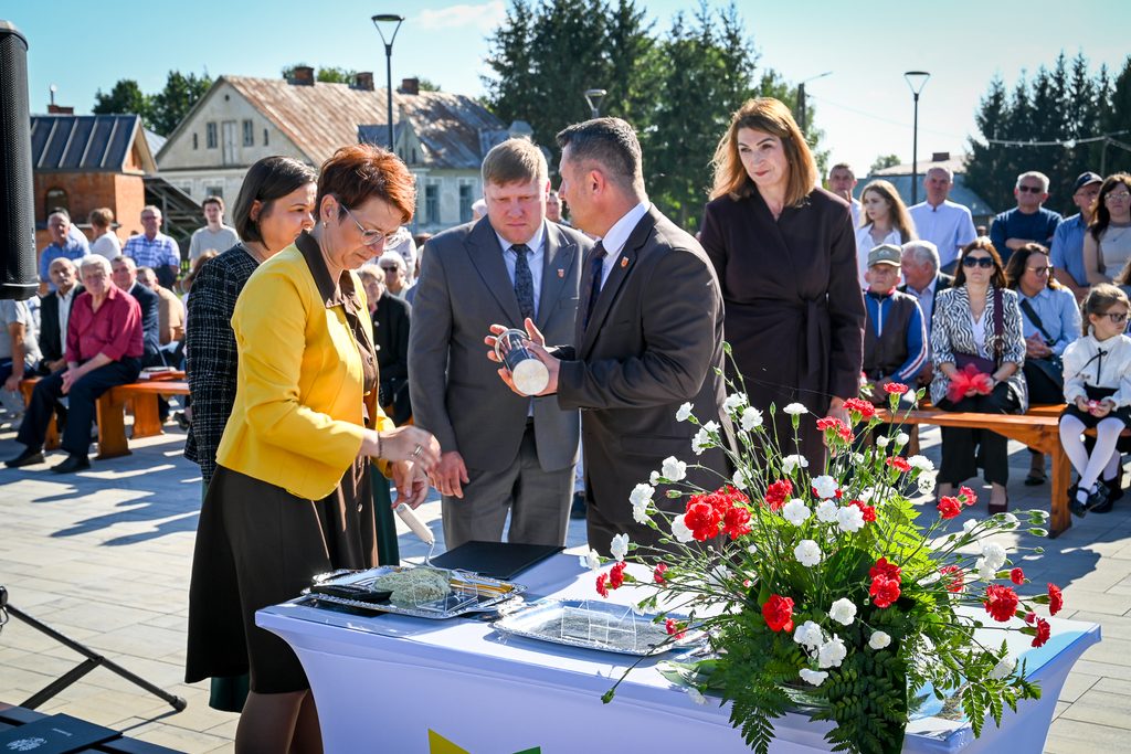 Grupa osób ubranych formalnie wokół stołu z białym obrusem, na którym stoją tace i kwiaty czerwone i białe, w tle ludzie na ławkach i domy.