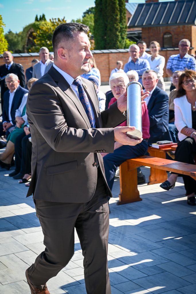 Mężczyzna w brązowym garniturze trzyma srebrną metalową tubę, w tle grupa ludzi siedzących na ławkach na zewnątrz, ceglane budynki i zieleń.