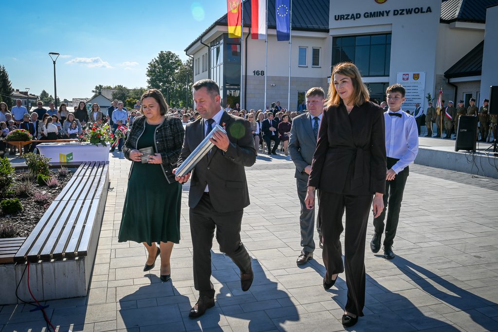 Cztery osoby idą na tle budynku Urzędu Gminy Dzwola, mężczyzna trzyma metalową tubę, kobiety w eleganckich ubraniach, w tle siedzący ludzie i żołnierze w mundurach.