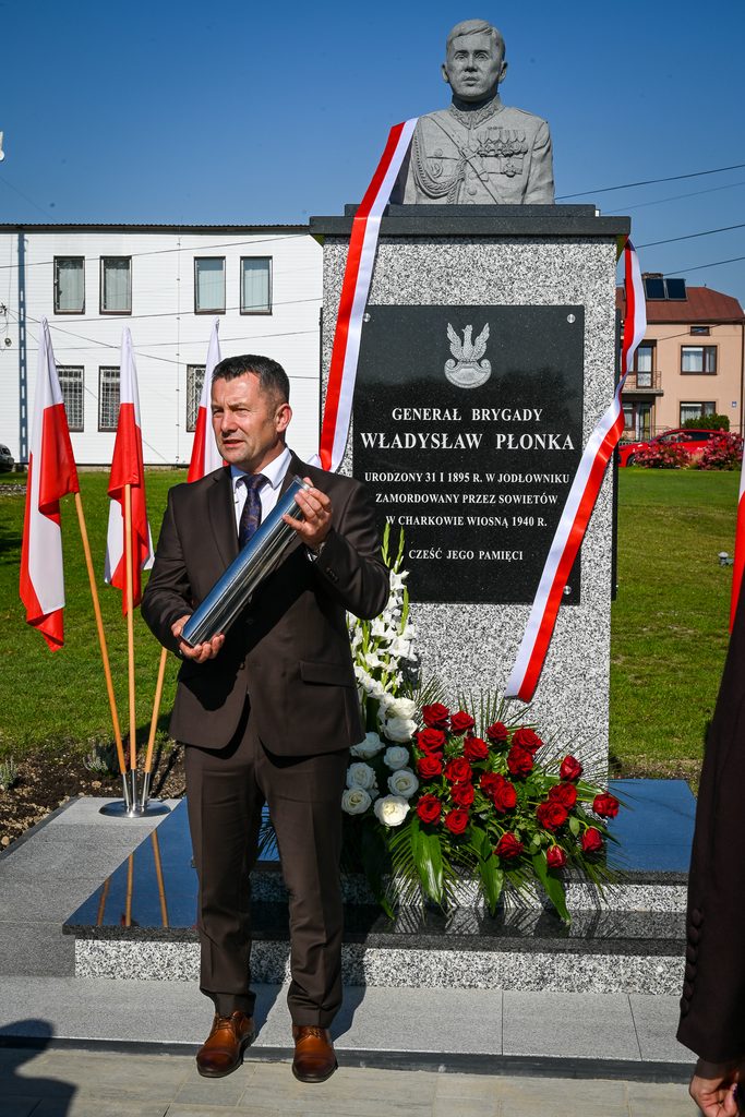 Pomnik generała Władysława Płonki z granitową tablicą i popiersiem, mężczyzna w ciemnym garniturze trzyma metalowy walec, obok biało-czerwone flagi i bukiet róż czerwonych i białych.