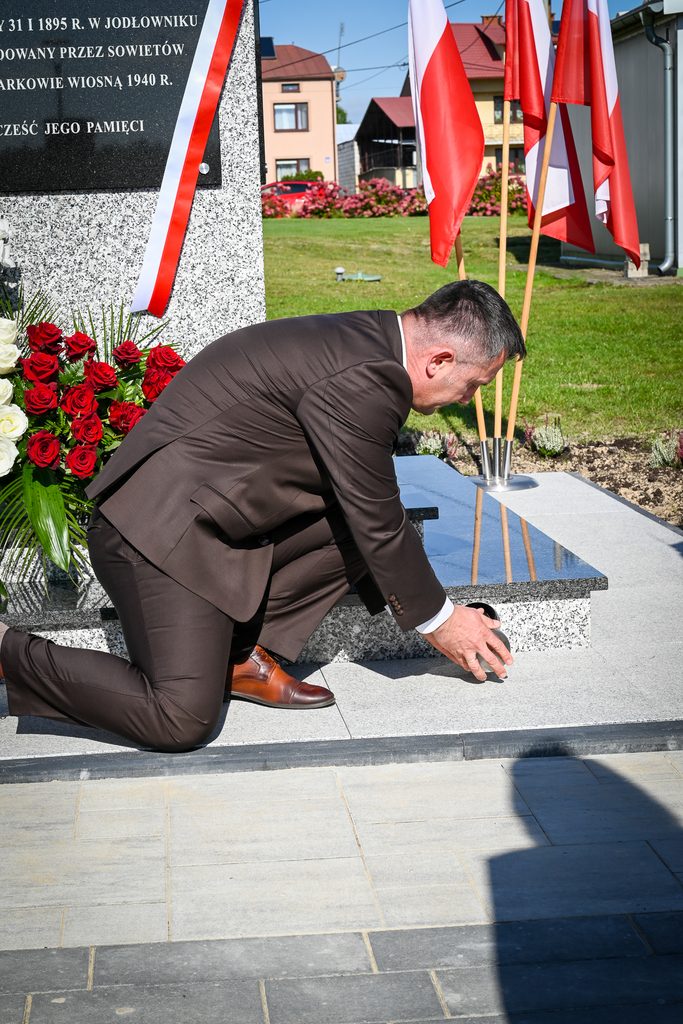 Mężczyzna w brązowym garniturze klęczy przy pomniku z czerwono-białą szarfą i bukietem czerwonych i białych róż, w tle polskie flagi na tle zielonej trawy i domów.