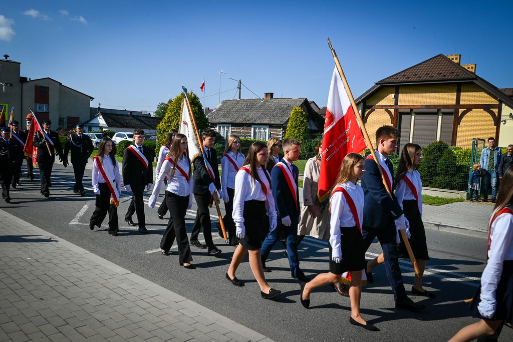 Młodzież w białych koszulach i czerwonych szarfach niesie flagi, idzie ulicą wzdłuż chodnika, w tle domy i kilka osób obserwujących przemarsz.