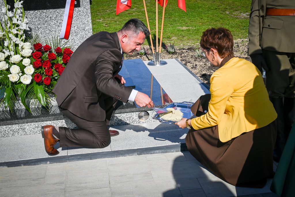 Mężczyzna w ciemnym garniturze i kobieta w żółtej kurtce przy granitowym pomniku z biało-czerwonymi kwiatami i flagami, kobieta trzyma tacę z mąką.