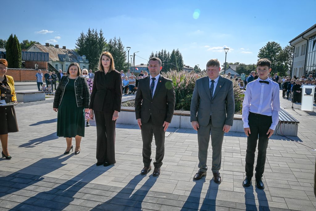 Grupa pięciu osób stojących na szarej kostce brukowej, w tle ludzie na ławkach, drzewa, budynki, słoneczna pogoda, niebieskie niebo.