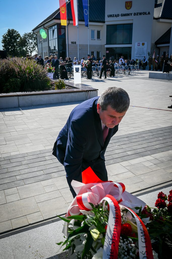 Mężczyzna w granatowym garniturze kładzie biało-czerwony wieniec z zielonymi liśćmi i kwiatami na tle placu przed budynkiem Urzędu Gminy Dzwola, w tle ludzie i flagi.