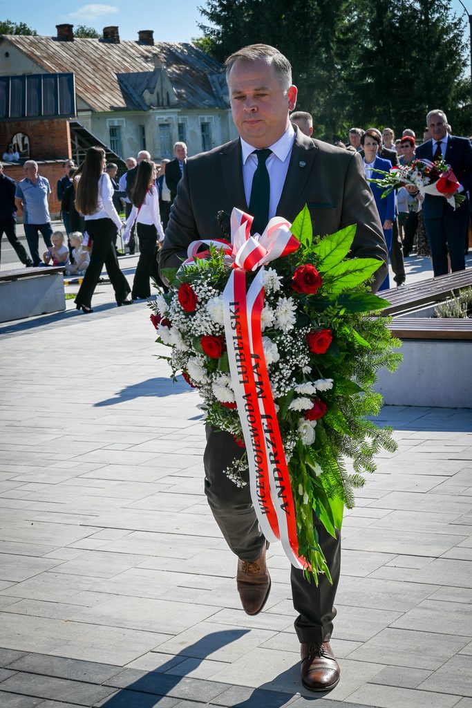Mężczyzna w ciemnym garniturze niesie wieniec z czerwonych i białych kwiatów z białą wstęgą z napisem, w tle grupa ludzi na otwartym placu, budynki i drzewa.