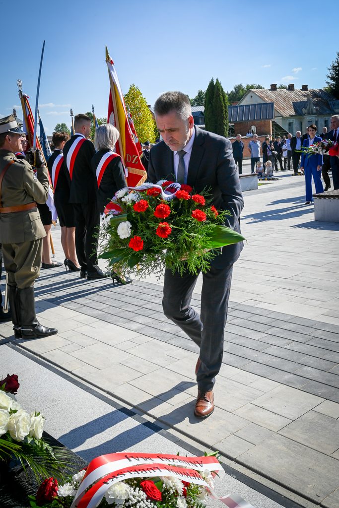 Mężczyzna w garniturze składający wieniec z czerwonych i białych kwiatów, obok osoby z mundurem wojskowym i grupy osób z flagami, na tle budynków i nieba.