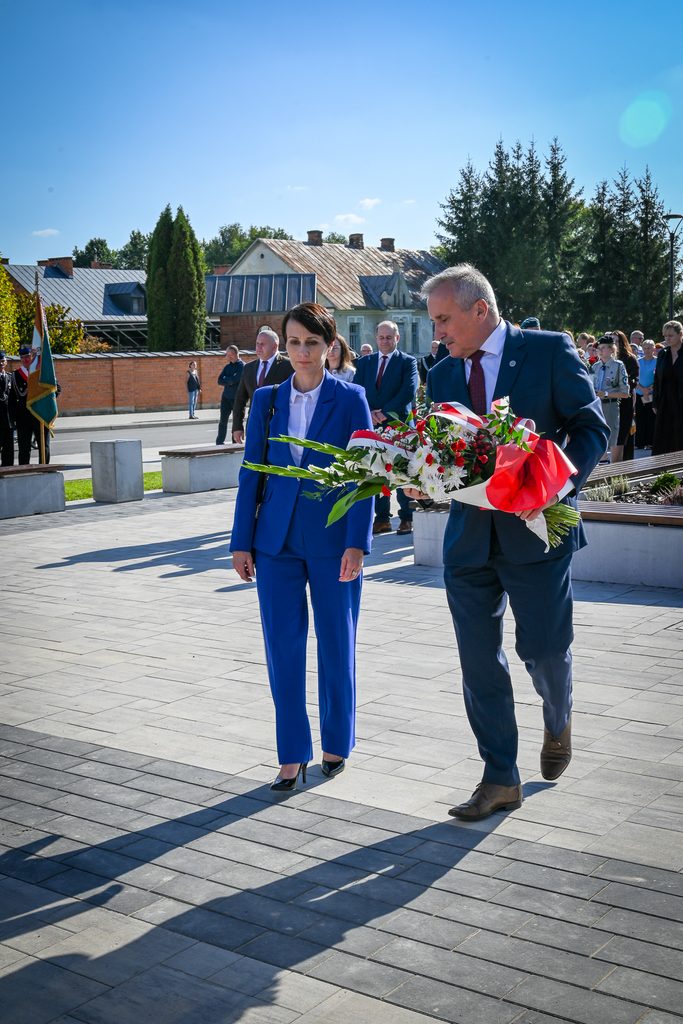 Mężczyzna w granatowym garniturze niesie bukiet kwiatów z białymi i czerwonymi kwiatami, obok kobieta w niebieskim garniturze, w tle ludzie i budynki pod błękitnym niebem.
