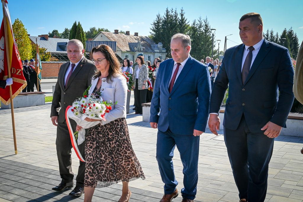 Grupa osób w garniturach i kobieta w jasnej marynarce z bukietem kwiatów z biało-czerwoną wstążką, idąca po brukowanej nawierzchni, w tle budynki, drzewa i flaga.