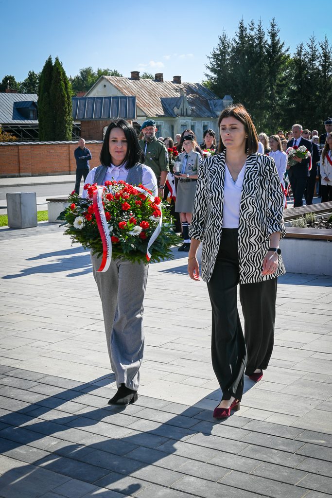 Grupa ludzi na zewnątrz, dwie kobiety na pierwszym planie niosąca wieniec z czerwonych i białych kwiatów, w tle drzewa, budynki i więcej osób w różnym wieku.