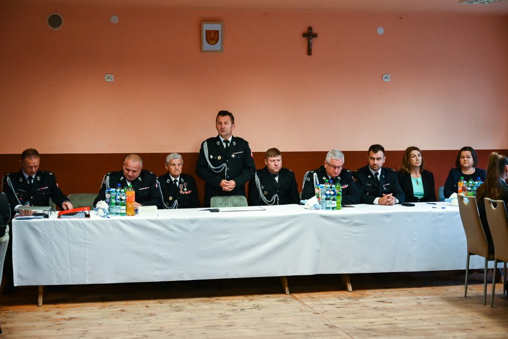 Dziesięć osób siedzących i jedna stojąca przy białym stole na tle pomarańczowej ściany z krzyżem i godłem, większość w mundurach czarnych z odznakami, na stole butelki i dokumenty.