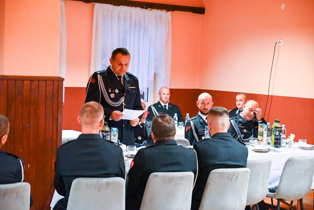 Grupa mężczyzn w mundurach siedzi przy stołach z białymi obrusami, jeden mężczyzna stoi i czyta z kartki, pomarańczowe ściany, białe zasłony, butelki i kartony napojów na stołach.