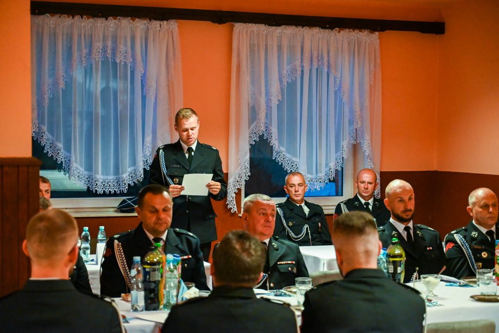 Mężczyźni w mundurach siedzą i stoją przy stołach z butelkami i szklankami, za nimi dwa okna z białymi koronkowymi firanami na pomarańczowej ścianie.