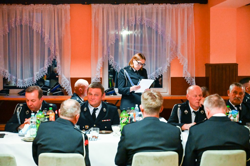 Grupa mężczyzn w mundurach siedzi przy stole z napojami, jedna kobieta stoi i czyta dokument, w tle pomarańczowe ściany i białe firanki z koronką na oknach.