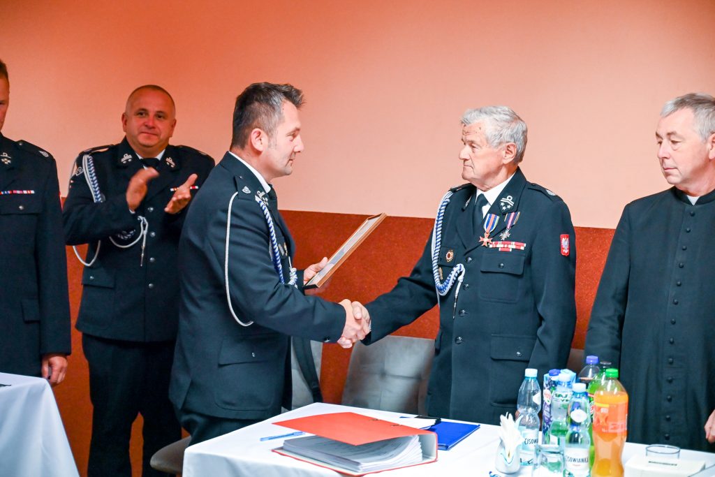 Mężczyźni w mundurach strażackich ściskają sobie dłonie, jeden trzyma ramkę, drugi ma medale, przy stole z dokumentami i napojami, na pomarańczowym tle.