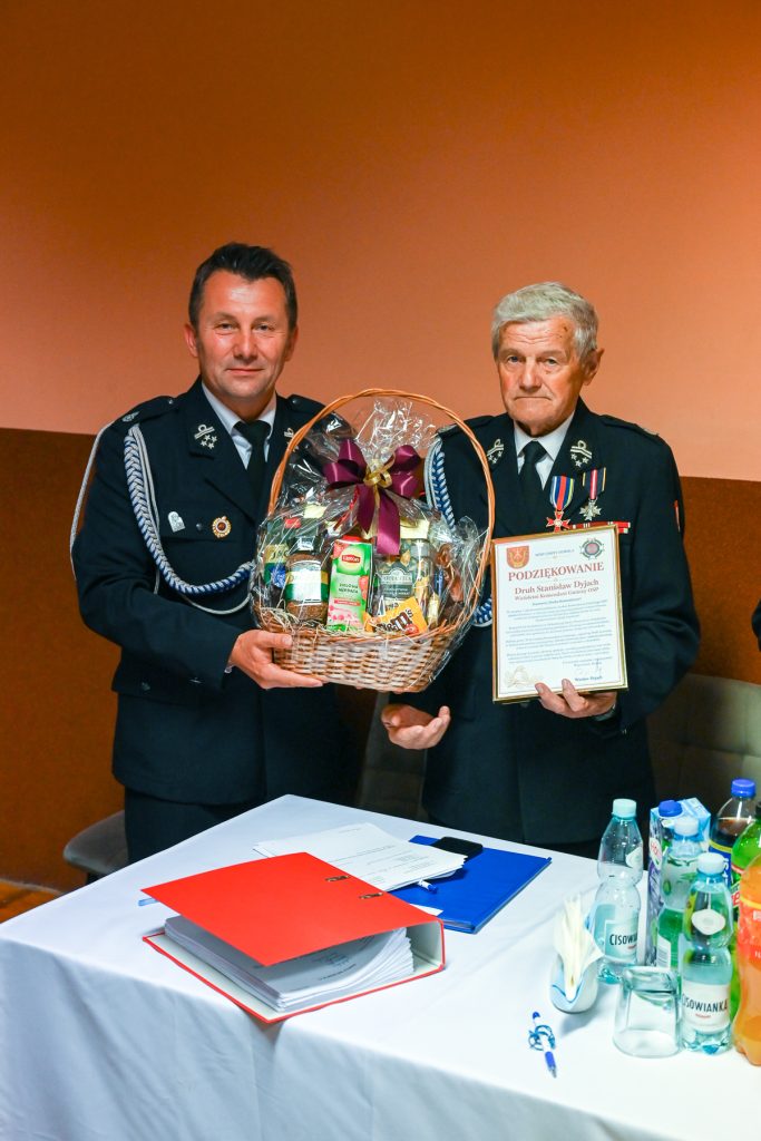 Dwaj mężczyźni w mundurach strażackich, jeden trzyma kosz upominkowy z kokardą, drugi dyplom podziękowania, biały stół z dokumentami i butelkami wody.