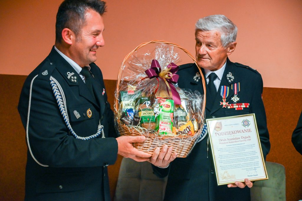 Dwaj mężczyźni w mundurach strażackich trzymają kosz wiklinowy z upominkami i dyplom podziękowania na tle pomarańczowej ściany.