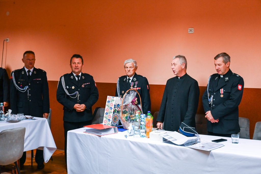 Pięciu mężczyzn w mundurach strażackich i dwóch w czarnych szatach duchownych stoi przy stole z białym obrusem, na stole koszyk prezentów i napoje, pomarańczowa ściana w tle.