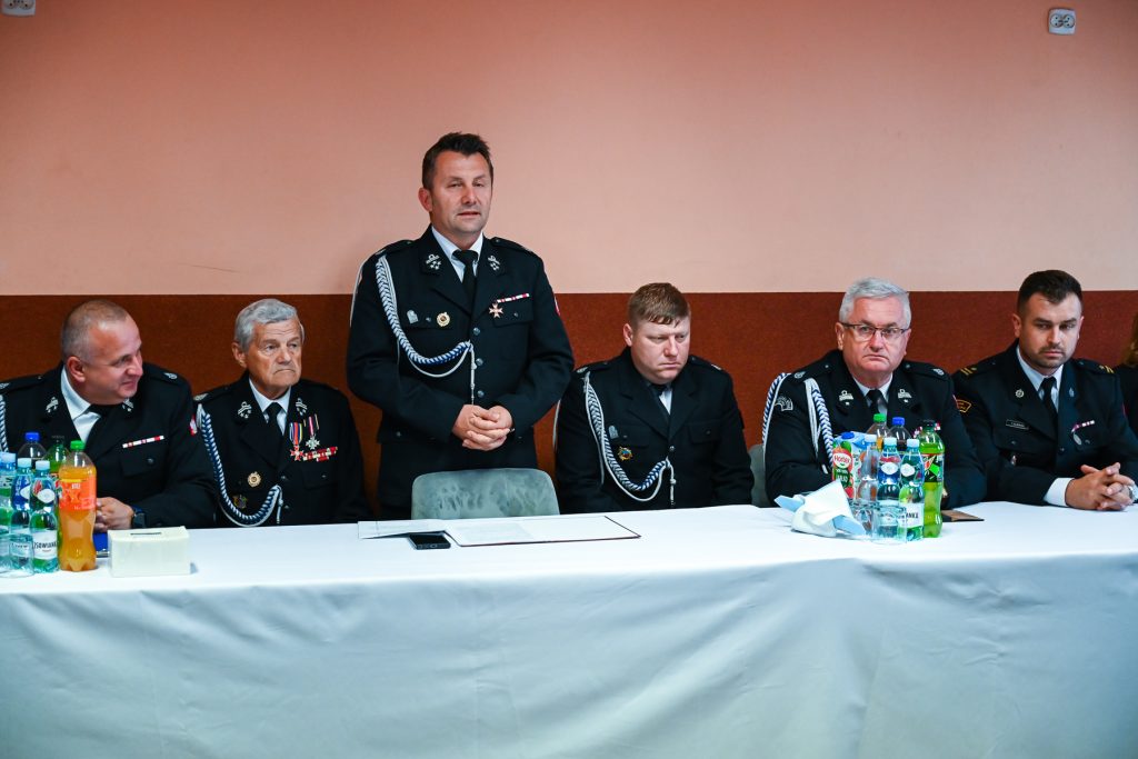 Sześciu mężczyzn w czarnych mundurach z odznaczeniami siedzi przy białym stole na tle pomarańczowej ściany, jeden stoi, na stole butelki z napojami i dokumenty.