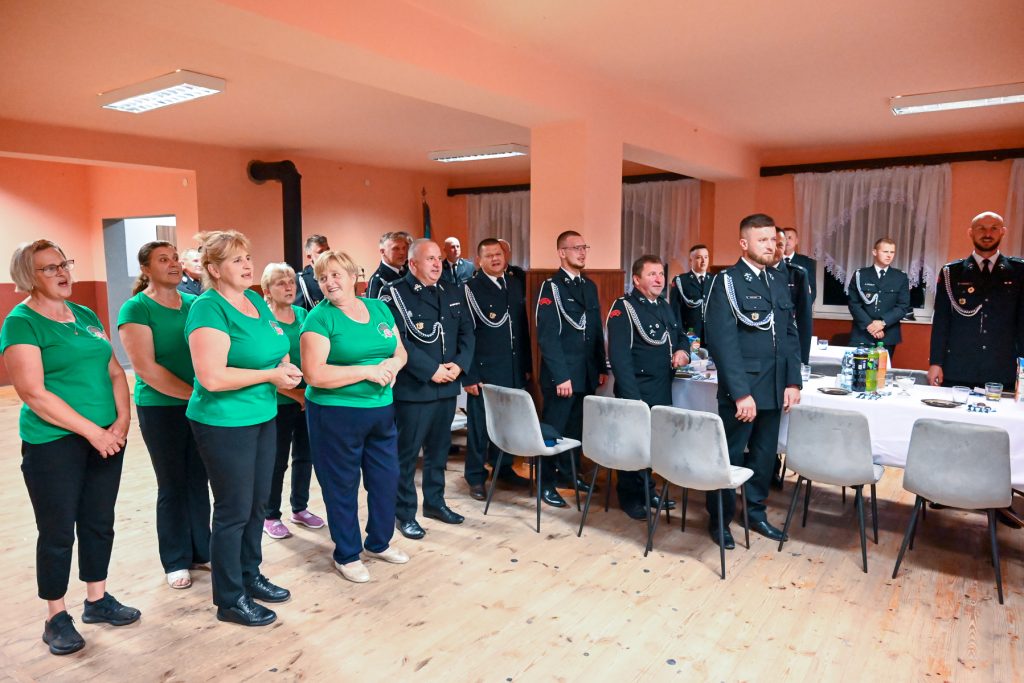 Grupa kobiet w zielonych koszulkach i mężczyzn w czarnych mundurach stojących w jasnym pomieszczeniu z drewnianą podłogą i stołem nakrytym białym obrusem.