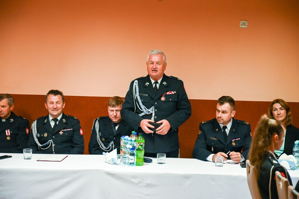 Mężczyzna w czarnym mundurze z odznaczeniami stoi za stołem, obok siedzą czterej mężczyźni i kobieta, wszyscy w mundurach, na stole butelki i szklanki, pomarańczowa ściana.