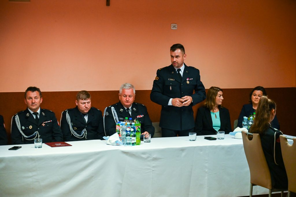 Mężczyźni w mundurach i kobiety w garniturach siedzą przy białym stole z napojami, na pomarańczowo-brązowej ścianie wisi krzyż.